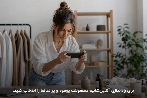 زنی جوان در حال عکس گرفتن از محصول روی میز چوبی با پس‌زمینه لباس‌های آویزان  و میز چوبی